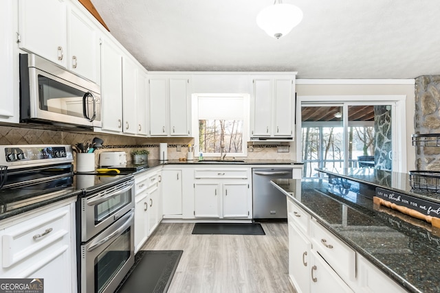 kitchen with appliances with stainless steel finishes, white cabinetry, dark stone counters, backsplash, and light hardwood / wood-style flooring