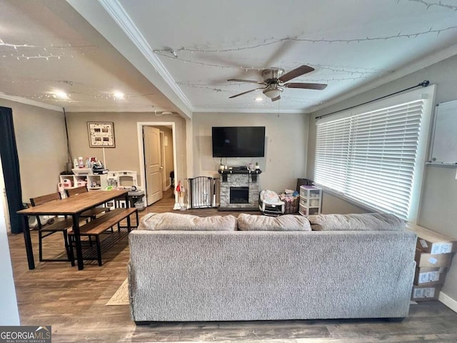 living room with ceiling fan, wood-type flooring, crown molding, and a stone fireplace