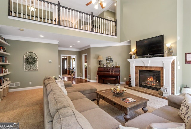 living room with a high ceiling, light carpet, ceiling fan, a brick fireplace, and crown molding