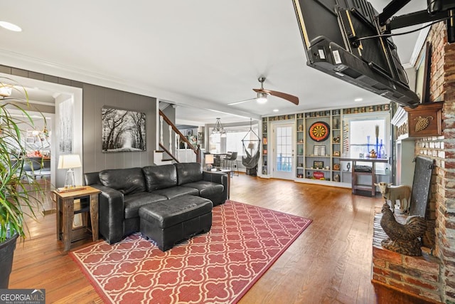 living room featuring hardwood / wood-style flooring, ornamental molding, ceiling fan with notable chandelier, and a brick fireplace