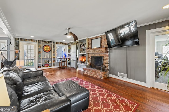 living room with ceiling fan, wood-type flooring, ornamental molding, a brick fireplace, and built in shelves