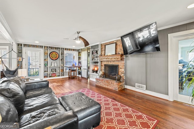 living room featuring built in features, ceiling fan, hardwood / wood-style floors, ornamental molding, and a brick fireplace