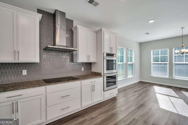 kitchen with decorative light fixtures, wall chimney range hood, dark stone countertops, white cabinetry, and double oven