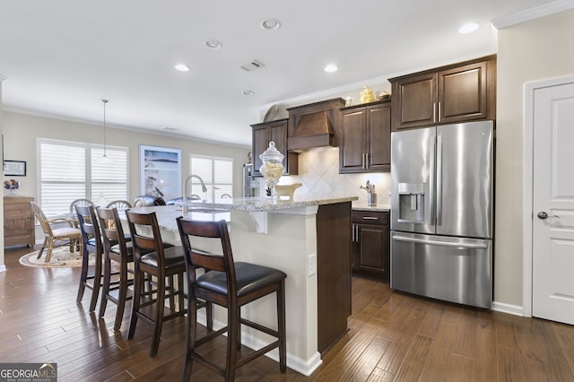 kitchen with premium range hood, dark wood-style flooring, visible vents, an island with sink, and stainless steel fridge