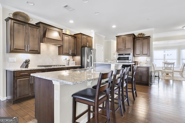 kitchen with premium range hood, dark wood-style flooring, visible vents, appliances with stainless steel finishes, and an island with sink