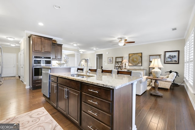 kitchen with an island with sink, dark wood-style floors, ornamental molding, double oven, and a sink