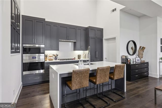 kitchen with a high ceiling, appliances with stainless steel finishes, a kitchen bar, dark hardwood / wood-style floors, and a center island with sink