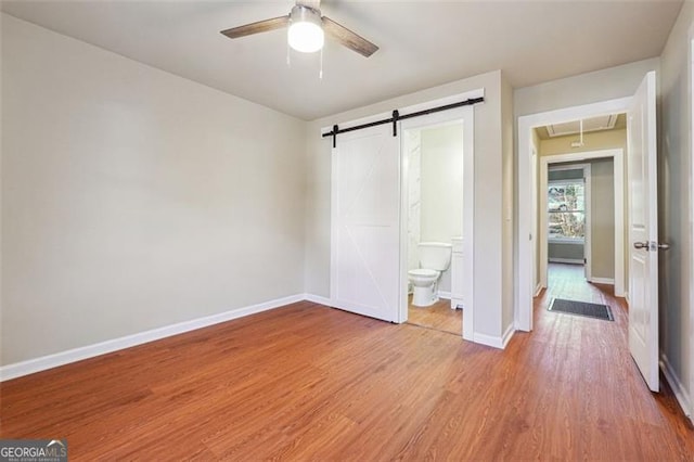 unfurnished bedroom with ceiling fan, a barn door, ensuite bathroom, and hardwood / wood-style flooring