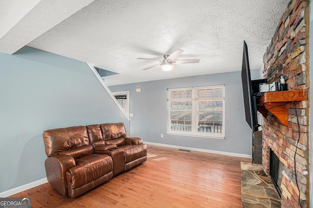 living room featuring a stone fireplace, a textured ceiling, ceiling fan, and light wood-type flooring