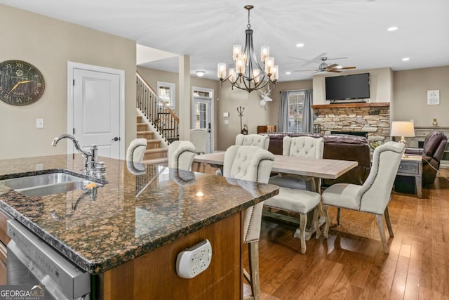 dining room with ceiling fan with notable chandelier, sink, a stone fireplace, and light wood-type flooring