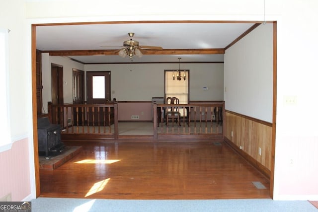 empty room featuring a wainscoted wall, ceiling fan with notable chandelier, wood finished floors, and a wood stove