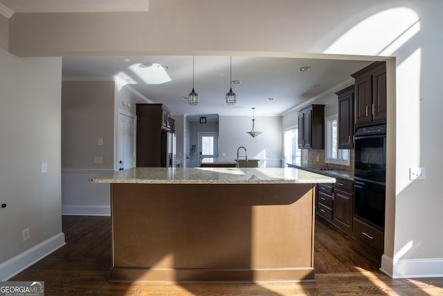 kitchen with pendant lighting, a center island with sink, dark brown cabinetry, double oven, and light stone counters