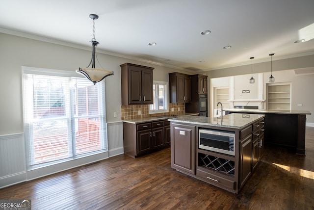 kitchen with stainless steel microwave, pendant lighting, sink, dark brown cabinetry, and an island with sink