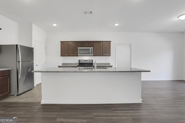 kitchen featuring dark brown cabinetry, stainless steel appliances, dark stone countertops, sink, and a kitchen island with sink