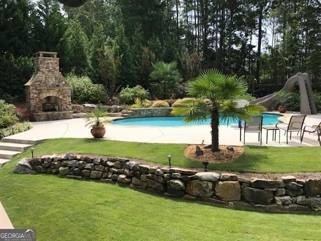 view of swimming pool featuring a patio, a water slide, an outdoor stone fireplace, and a lawn