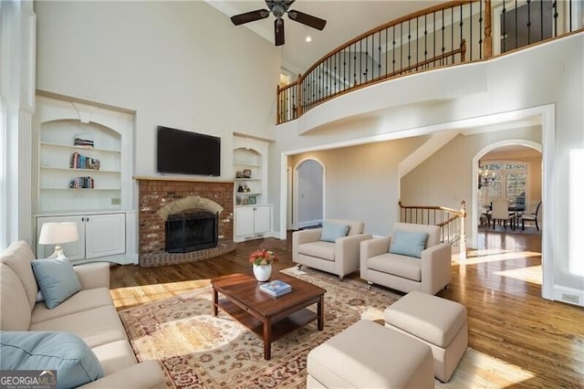 living room featuring built in shelves, a towering ceiling, ceiling fan, a fireplace, and light hardwood / wood-style floors