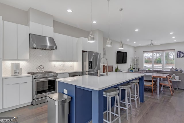 kitchen featuring wall chimney range hood, a center island with sink, high end appliances, hanging light fixtures, and white cabinets