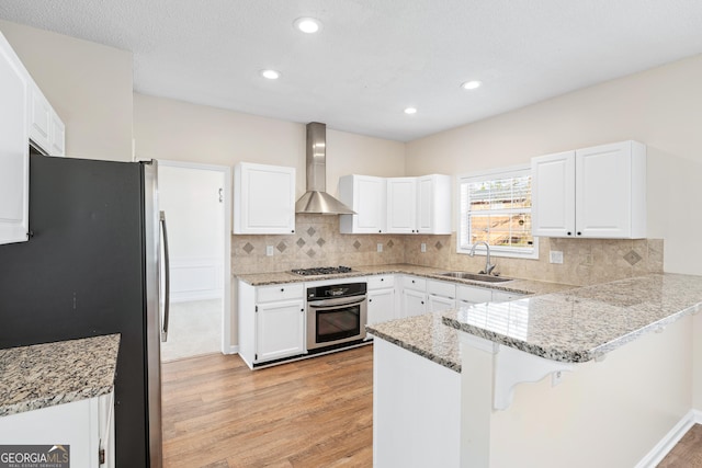 kitchen featuring white cabinets, appliances with stainless steel finishes, wall chimney range hood, sink, and kitchen peninsula