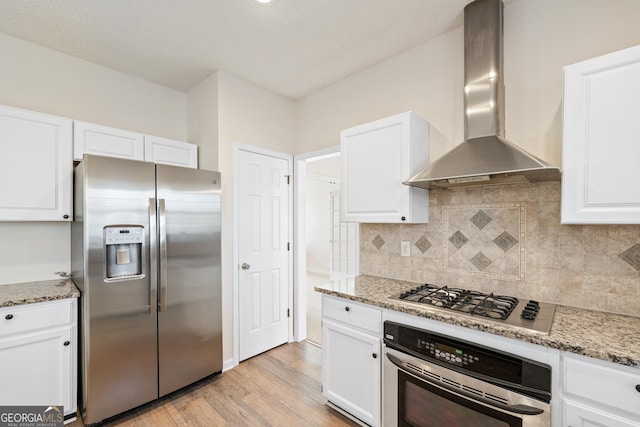 kitchen with wall chimney range hood, stone counters, light hardwood / wood-style floors, white cabinetry, and appliances with stainless steel finishes