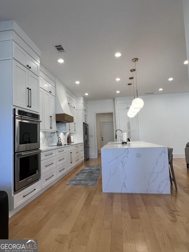 kitchen with custom exhaust hood, white cabinets, stainless steel appliances, and a center island with sink