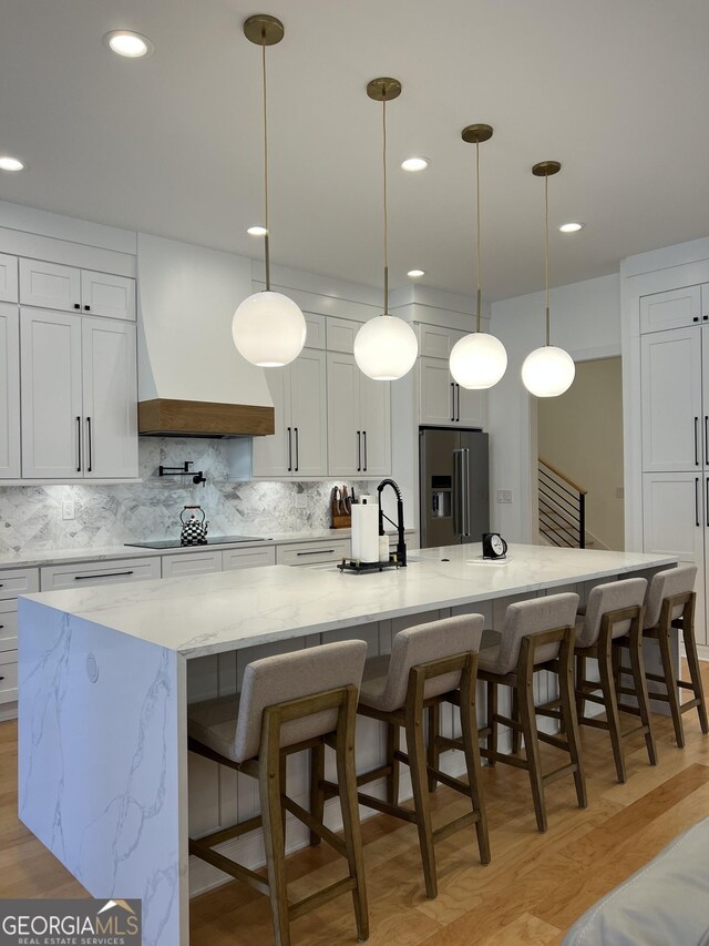 kitchen with a large island with sink, black electric cooktop, high end refrigerator, and custom range hood