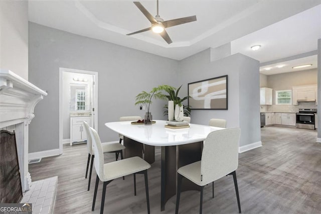 dining area featuring a brick fireplace, a tray ceiling, ceiling fan, and light wood-type flooring