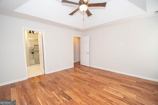 unfurnished bedroom with ceiling fan, light hardwood / wood-style floors, a tray ceiling, and ensuite bath