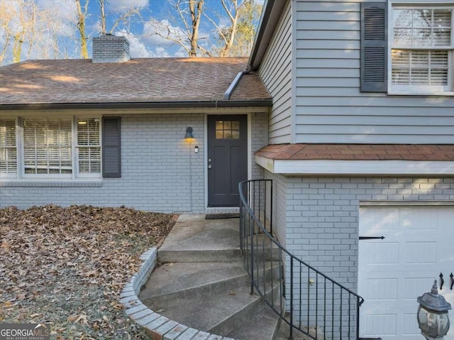 doorway to property with a garage, brick siding, a shingled roof, and a chimney