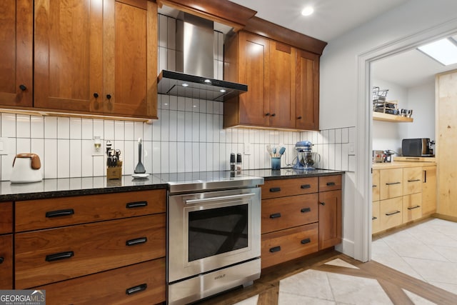 kitchen featuring wall chimney range hood, dark stone counters, backsplash, stainless steel range with electric stovetop, and light tile patterned flooring