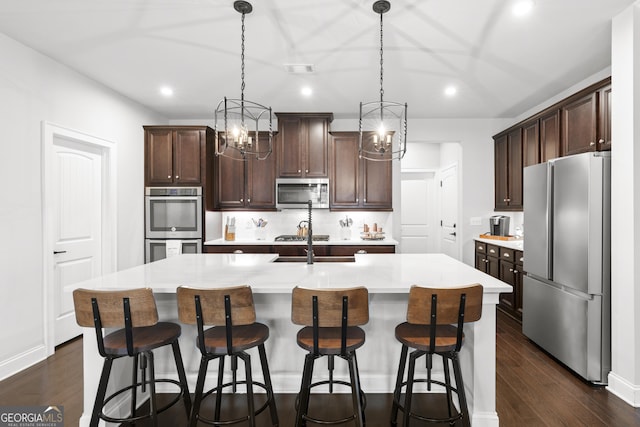 kitchen with appliances with stainless steel finishes, pendant lighting, dark hardwood / wood-style flooring, dark brown cabinetry, and a kitchen island with sink