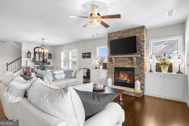 living area with visible vents, stairs, hardwood / wood-style floors, a stone fireplace, and ceiling fan with notable chandelier