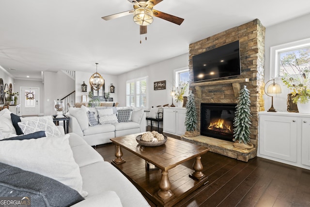 living room featuring ceiling fan with notable chandelier, dark wood-type flooring, and a stone fireplace