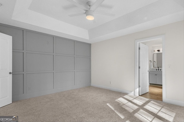 unfurnished bedroom with ceiling fan, light colored carpet, a tray ceiling, and ensuite bathroom