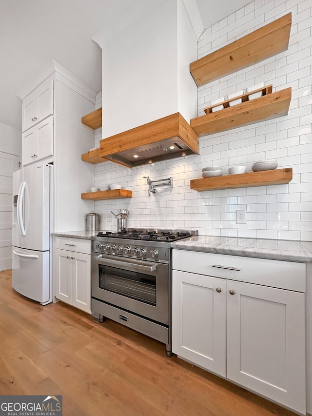 kitchen featuring backsplash, stainless steel range, white refrigerator with ice dispenser, white cabinets, and light stone counters