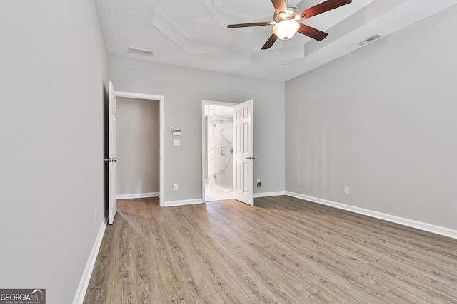 unfurnished bedroom featuring a textured ceiling, connected bathroom, light hardwood / wood-style floors, a raised ceiling, and ceiling fan