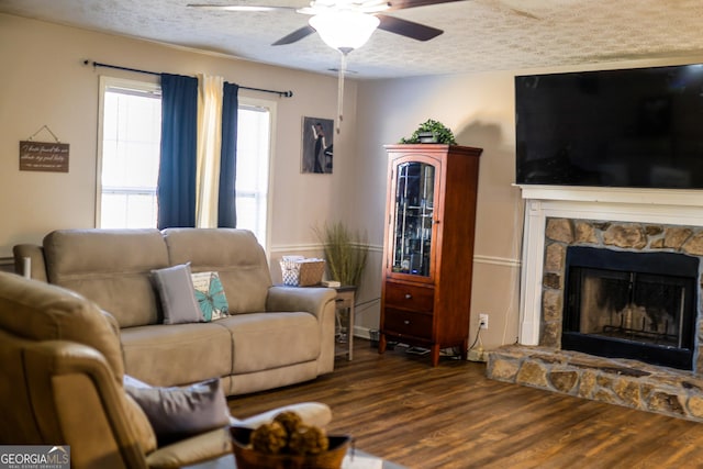 living room featuring ceiling fan, a textured ceiling, dark hardwood / wood-style floors, and a stone fireplace