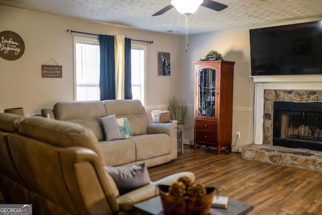 living room with ceiling fan, a stone fireplace, a textured ceiling, and hardwood / wood-style flooring