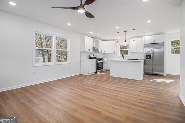 kitchen featuring wall chimney range hood, white cabinetry, stainless steel appliances, decorative light fixtures, and light wood-type flooring