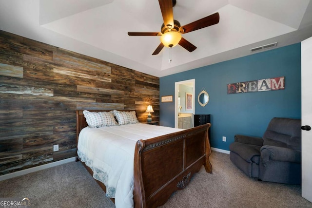 carpeted bedroom with ceiling fan, a tray ceiling, wooden walls, and ensuite bath