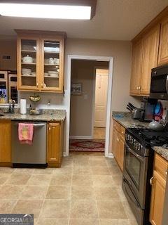 kitchen featuring appliances with stainless steel finishes, brown cabinetry, glass insert cabinets, and light stone counters