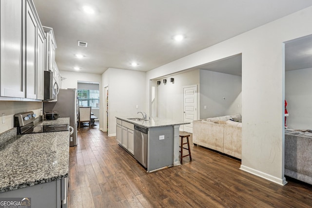 kitchen with appliances with stainless steel finishes, dark wood-type flooring, a kitchen island with sink, light stone counters, and a breakfast bar