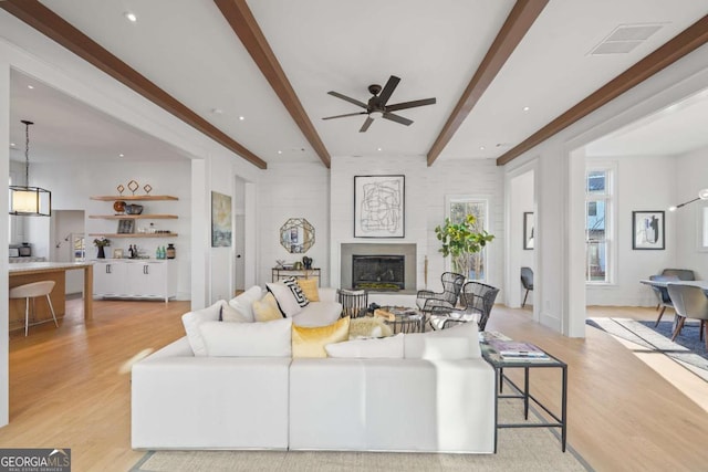 living room with visible vents, a ceiling fan, light wood-type flooring, beamed ceiling, and a glass covered fireplace