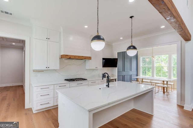 kitchen with white cabinets, premium range hood, backsplash, light stone counters, and a center island with sink