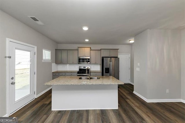 kitchen featuring stainless steel appliances, an island with sink, gray cabinetry, and light stone counters