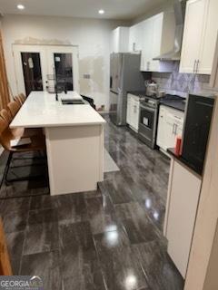 kitchen with wall chimney exhaust hood, white cabinetry, stainless steel appliances, a kitchen island with sink, and a breakfast bar area