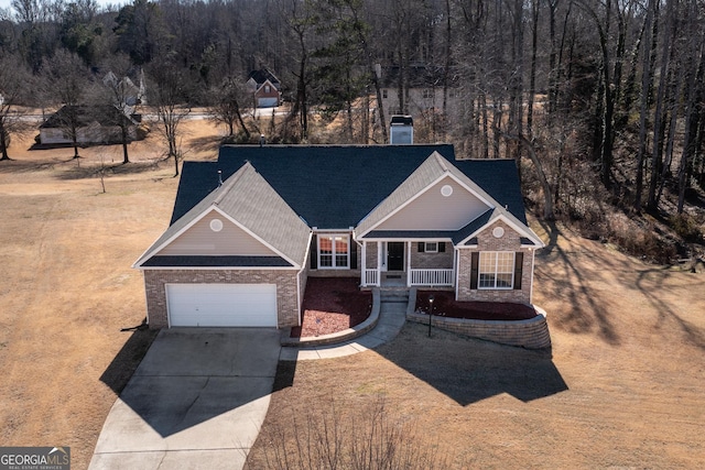 view of front of property featuring brick siding, covered porch, an attached garage, a wooded view, and driveway