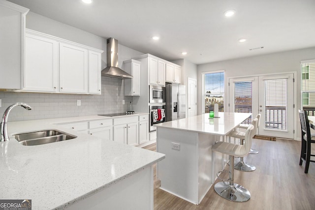 kitchen with wall chimney range hood, a kitchen island, sink, stainless steel appliances, and white cabinets