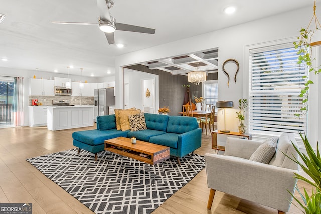 living room featuring ceiling fan with notable chandelier, light wood-type flooring, beamed ceiling, and coffered ceiling