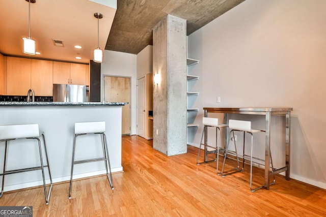 kitchen with a kitchen bar, dark stone countertops, hanging light fixtures, stainless steel refrigerator, and light wood-type flooring