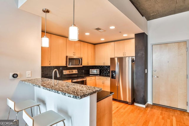 kitchen featuring decorative light fixtures, kitchen peninsula, stainless steel appliances, light brown cabinetry, and dark stone counters